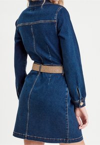 Robe en denim bleu marine, dotée de manches longues, d'une taille ceinturée et de boutons décoratifs aux poignets. Texture lisse avec des détails de couture visibles.