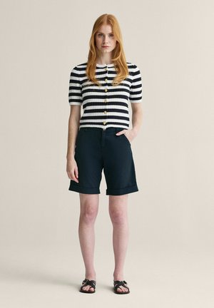 Ung kvinde med rødt hår iført en sort og hvid stribet kortærmet knapbluse, sorte knælange shorts og sorte sandaler, stående neutralt.