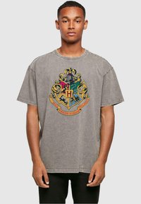 Camiseta gris de gran tamaño con un emblema vibrante y multicolor que presenta un escudo y una pancarta con texto, fabricada en suave material de algodón.