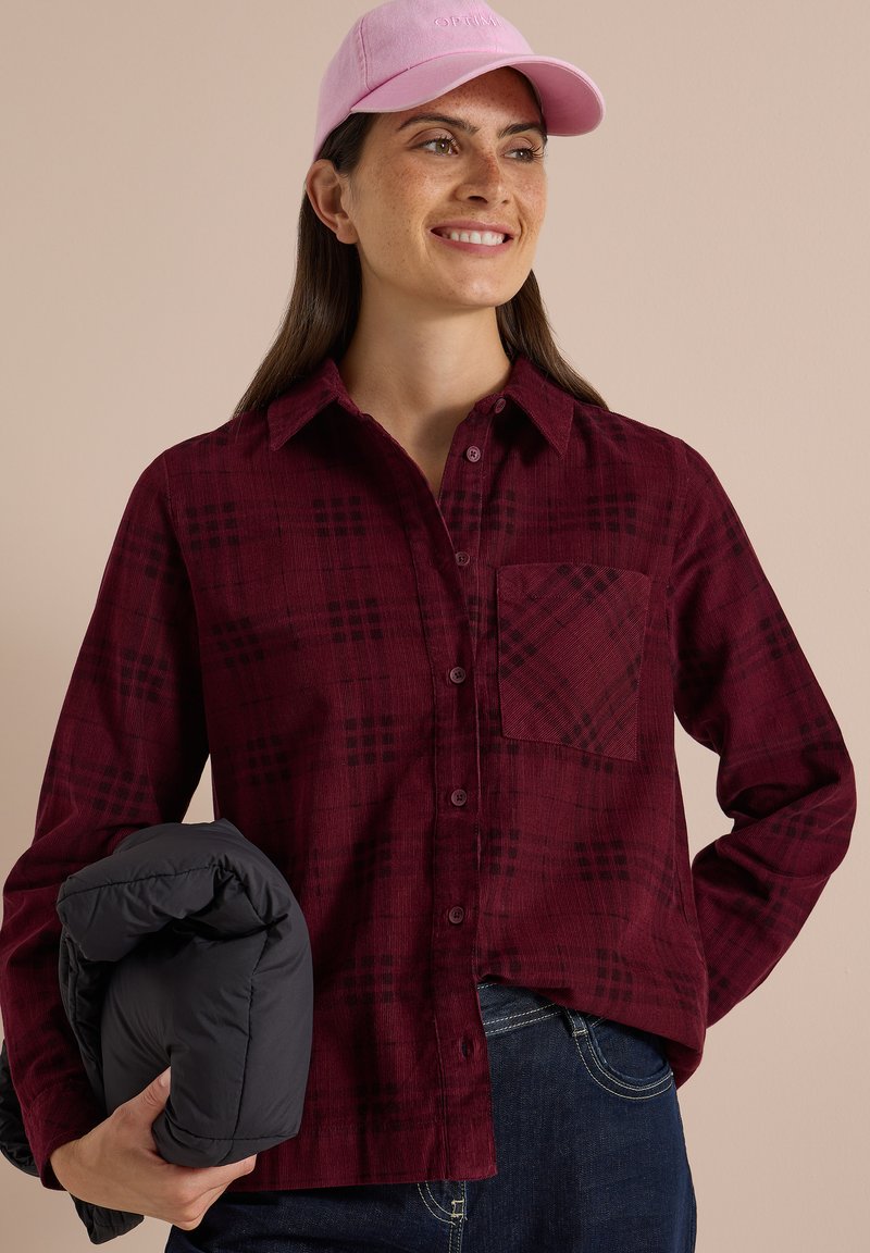Femme portant une casquette rose et une chemise à carreaux bordeaux, souriante et tenant une veste noire pliée dans sa main gauche.