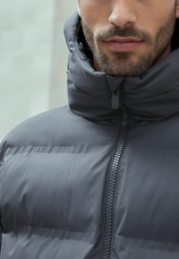 Tamsiai pilkas pūkinis švarkas, su aukštu kaklu ir pilnu priekiu užtrauktuku su tekstūruotu užtrauktuku. Lygi audinio struktūra su siuvinėtų sekcijų.