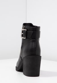 Bottine en cuir noir avec un talon épais, fermeture éclair sur le côté et une sangle avec une boucle en argent. Texture lisse et design à bout rond.