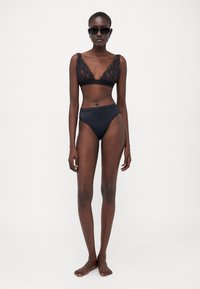 Bralette di pizzo nera con spalline larghe, abbinato a slip bikini neri a vita alta. La modella indossa occhiali da sole scuri e è a piedi nudi.