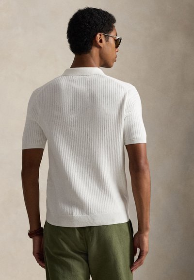 Ribbed short-sleeve polo shirt in white, featuring a classic collar, textured knit design, and a relaxed fit, paired with green trousers.