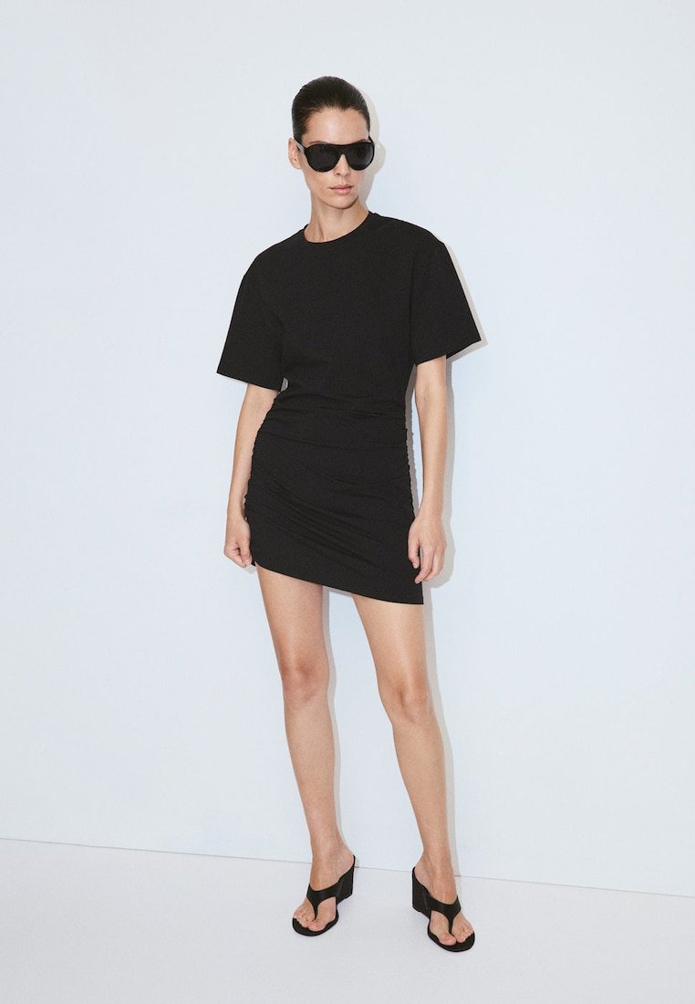 Femme portant une robe noire oversize à manches courtes, des sandales noires à talons hauts et de grandes lunettes de soleil noires, debout contre un mur blanc uni.