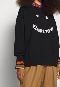 Sudadera negra con un gran gráfico de texto sonriente en blanco, con puños de colores a rayas asomándose, de corte holgado.