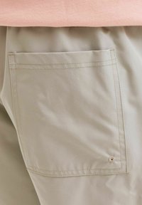 Τσέπη από μπεζ υφασμάτινο παντελόνι με μπεζ ραφές και μικρή στρογγυλή κεντημένη λεπτομέρεια κοντά στην κάτω γωνία, κάτω από ένα ροζ ανοιχτό ρούχο.