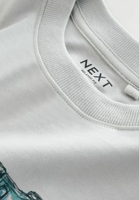 Ljust grå bomullst-shirt med ribbad rund hals och synlig varumärkesetikett "NEXT" med avslappnad passform. Slät textur.