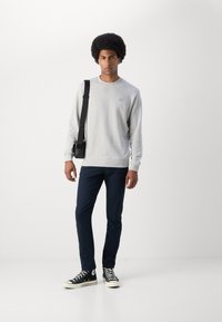Gray sweatshirt with a round neck, black accent logo on chest, paired with navy pants and black high-top sneakers. Model holds a black messenger bag.