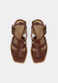 Καφέ δερμάτινα σανδάλια με πολλαπλούς ιμάντες, τετράγωνη μύτη και ιμάντα αστραγάλου με πόρπη. Λείο υλικό με επίπεδη σόλα.