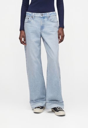 Persoon draagt lichtblauwe wijde jeans met opgerolde boorden, zwart-witte sneakers en een marineblauwe longsleeve, staand tegen een witte achtergrond.