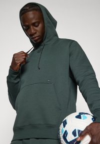 Felpa verde con tasca a marsupio e logo Nike. Presenta un cappuccio con coulisse e polsini a costine. Tiene un pallone da calcio.