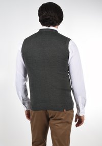 Mørkegrøn strikket vest med ribbet kant og krave, båret over en hvid skjorte, med et lille logo-tag på den nederste side.