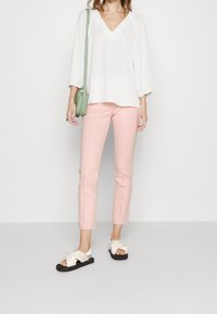 Vit V-ringad blus med ballongärmar, kombinerad med ljusrosa skräddarsydda byxor. Vita sandaler med korsremmar på svart sula. Grön axelväska.