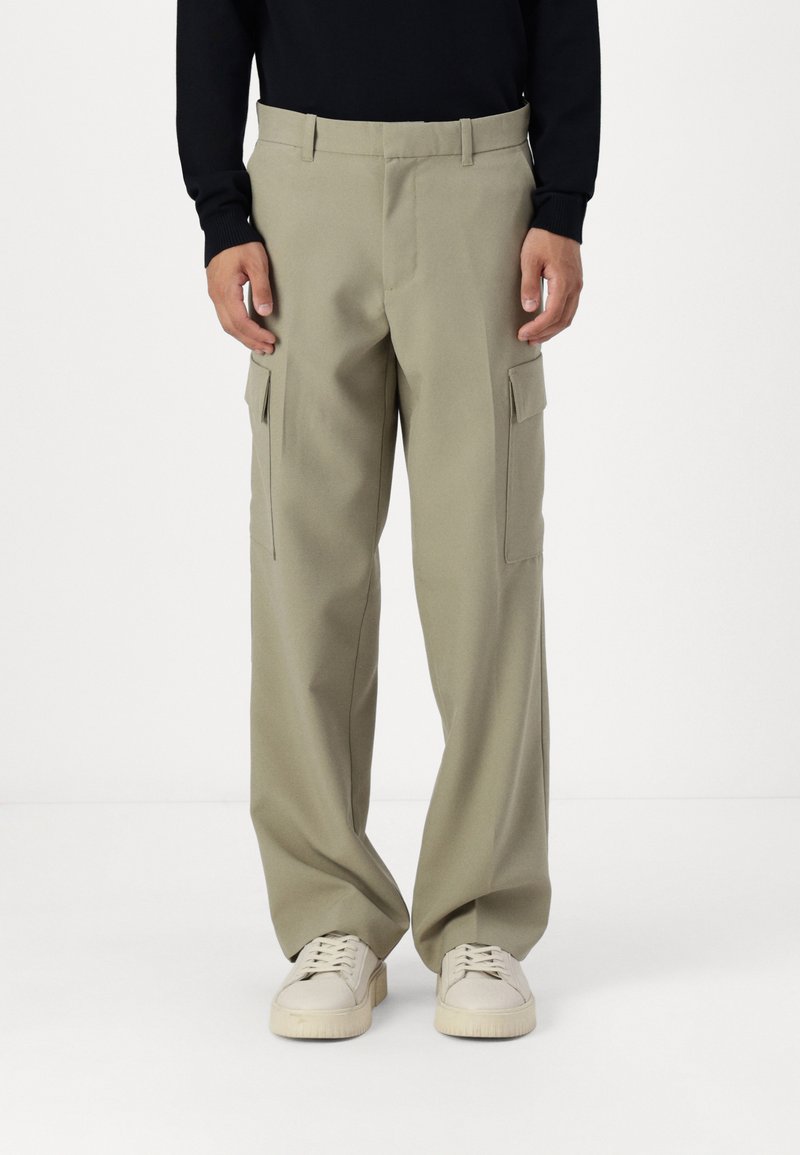 Pantalones cargo color caqui de corte holgado, con dos bolsillos laterales y una textura suave. Combinados con zapatillas ligeras sobre un fondo neutro.