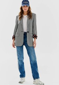 Sivý houndstooth blejzer s jedným gombíkom, kombinovaný s modrými džínsami so bočnými pruhmi a bielymi teniskami. Modrá baseballová čiapka s textom.