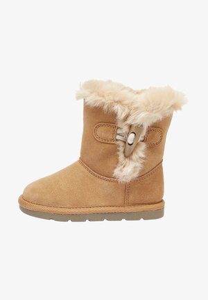 Botte d’hiver en daim beige avec doublure en fourrure duveteuse et bouton décoratif sur le côté, semelle en caoutchouc plate et hauteur mi-mollet.