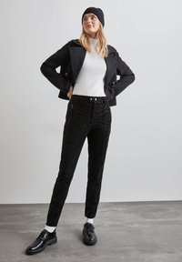 Veste noire à double boutonnage, col roulé blanc à côtes, pantalon noir à imprimé léopard avec zips sur les côtés, chaussures noires et bonnet en tricot noir.
