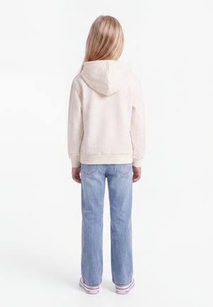 Enfant aux longs cheveux blonds portant un sweat à capuche crème, un jean bleu clair et des baskets blanches, debout de dos sur un fond blanc.
