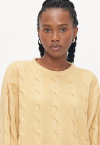Pull en maille torsadée de couleur jaune doux. Avec un col ras du cou et des manches longues. Le tissu présente une finition texturée avec une coupe confortable et décontractée.