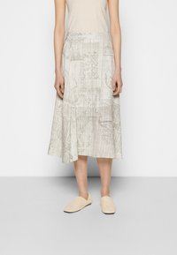Vaaleanbeige midi-hame, jossa on teksturoitu ja kuvioitu pinta, hienovaraisia kuvioita ja A-linjainen siluetti. Yhdistettynä vaaleanbeigeihin slip-on-kenkiin.