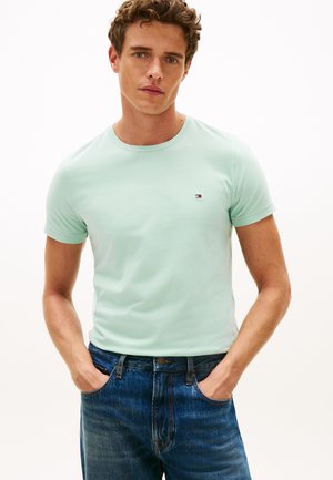 Jeune homme aux cheveux bouclés portant un t-shirt vert clair à manches courtes et un jean bleu, debout les mains dans les poches devant un fond blanc uni.