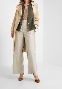 Manteau trench beige, cardigan vert olive, débardeur beige, pantalon en lin à jambes larges et talons rayés marron. Détails notables incluent les boutons et la coupe.