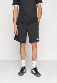 Γκρι αθλητικό σορτς με τρεις λευκές διαγώνιες ρίγες και υφασμάτινη υφή, σε συνδυασμό με μαύρο κοντομάνικο πουκάμισο και μαύρα αθλητικά παπούτσια.