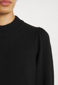 Svart stickad tröja med ribbat mönster, rund halsringning och puffiga axlar, med en mjuk finish och strukturerad design.