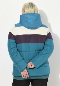 Giacca imbottita in teal, con un grande cappuccio, strisce orizzontali beige e viola scuro. Tessuto liscio con polsini elasticizzati e orlo a coste.