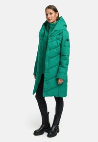 Zielony kurtka puchowa z kapturem, z pikowanym wzorem, zatrzaskami i kieszeniami. Noszona na czarną bluzkę i połączona z czarnymi spodniami oraz butami.