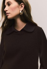 Brown knitted vest with ribbed edges over a black collared shirt. The model wears decorative gold earrings, showcasing a textured design.