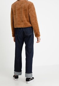 Giacca in pelliccia marrone, design corto, esterno testurizzato, abbinata a jeans di denim scuri. I jeans presentano orli arrotolati e una vestibilità rilassata. Sneakers nere visibili.