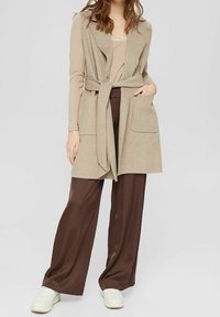 Gilet lavorato a maglia beige con cintura, due tasche, abbinato a pantaloni in satin marroni e sneakers bianche. Design semplice e texture morbida.