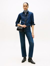 Vrouw staat rechtop en draagt een donkerdenim shirt en jeans, zwarte enkellaarsjes, een gestreepte coltrui, een kleine zwarte schoudertas en een gouden armband.