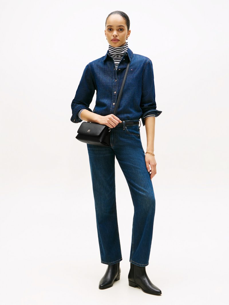 Vrouw staat rechtop en draagt een donkerdenim shirt en jeans, zwarte enkellaarsjes, een gestreepte coltrui, een kleine zwarte schoudertas en een gouden armband.
