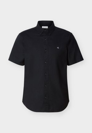 Zwarte kortärmelige blouse met knopen, kraag en klein wit "CK"-logo op de linkerborst, tegen een effen achtergrond.