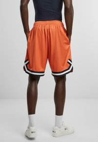 Orange mesh sportsbukser med sort og hvidt stribet kant, der har en elastisk talje og sidelommer.