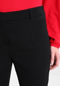 Zwarte op maat gemaakte broek met een gladde textuur, slim fit en een vlakke voorkant. Voorzien van een tailleband met riemlussen. Rode top gedeeltelijk zichtbaar.