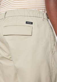 Pantaloni beige con elastico in vita, dotati di una tasca posteriore e di un'etichetta nera Calvin Klein. Tessuto morbido con una superficie liscia.