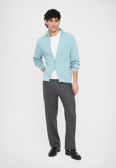 Ελαφρώς μπλε πουλόβερ με φερμουάρ και ριμπ ύφανση, φορεμένο πάνω από λευκό μπλουζάκι, σε συνδυασμό με σκούρο γκρι παντελόνι και μαύρα παπούτσια. Απλός, μοντέρνος σχεδιασμός.