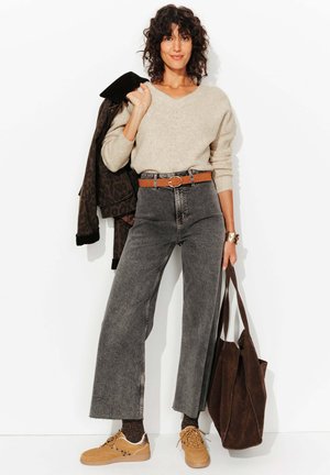 Un pull en tricot beige clair, un jean noir taille haute, une ceinture marron, des baskets beige, et un sac fourre-tout marron foncé. Cheveux bouclés, tenant une veste.