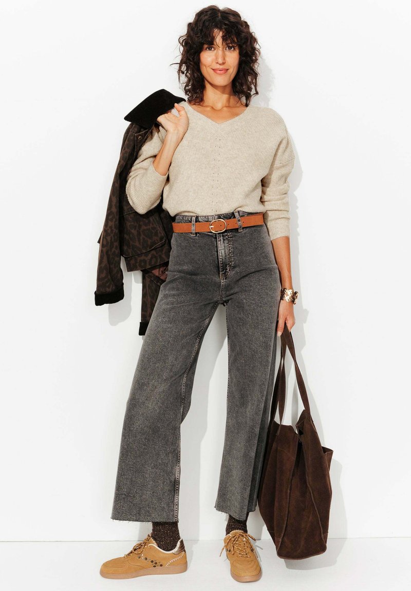 Un pull en tricot beige clair, un jean noir taille haute, une ceinture marron, des baskets beige, et un sac fourre-tout marron foncé. Cheveux bouclés, tenant une veste.