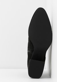 Botte noire à bout pointu avec semelle en caoutchouc et petit talon bloc, semelle face vers l'avant sur fond blanc.
