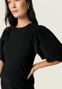 Schwarzes Kleid mit puffäugigen kurzen Ärmeln, rundem Ausschnitt und dezenten Seitennähten, gefertigt aus einem glatten, leicht strukturierten Stoff.
