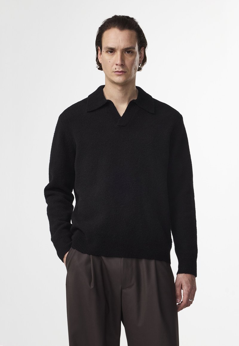 Maglione nero a texture con colletto ampio e scollo a V. La vestibilità è rilassata, abbinato a pantaloni marroni larghi.