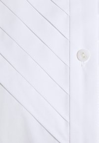 Primo piano della parte anteriore di una camicia bianca con pieghe diagonali e un bottone bianco sul lato destro.