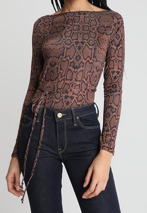 Nainen, jolla on päällään pitkähihainen, ruskea käärmekuvioinen istuva paita sivusolmulla, yhdistettynä tummansinisiin korkeavyötäröisiin farkkuihin.