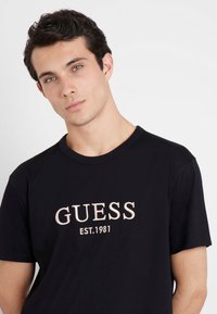 T-shirt en coton noir avec un logo brodé beige bien visible, manches courtes et col rond.