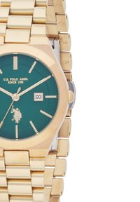 Montre-bracelet en ton doré avec un cadran vert, des accents argentés, affichage de la date et un bracelet en métal maillé. Présente un logo de joueur de polo et des marqueurs d'heure.